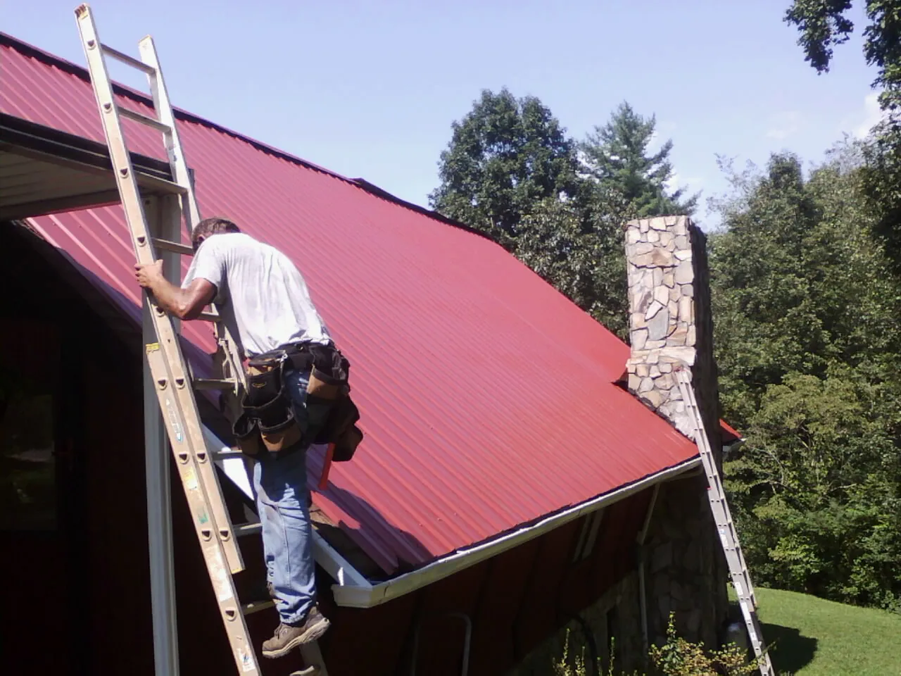Roofer on a ladder beside a red metal roof and stone chimney — Soffit & Fascia Repair by Roofing Brothers in Montevideo